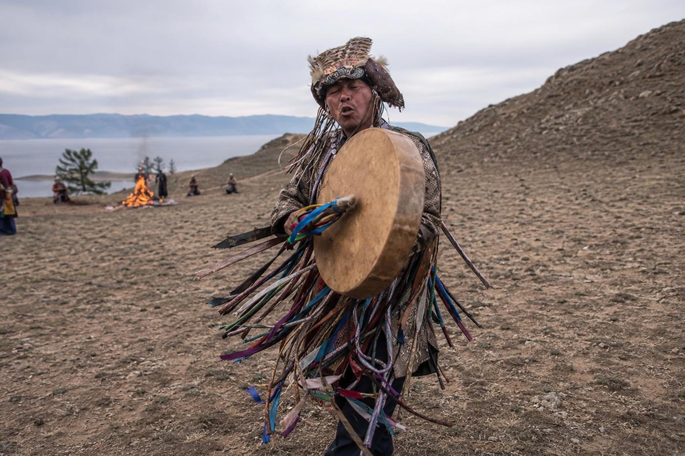 A shaman in a ribboned ritual coat and feathered headdress striking his drum on the open steppe, the lake and a ceremonial fire behind him