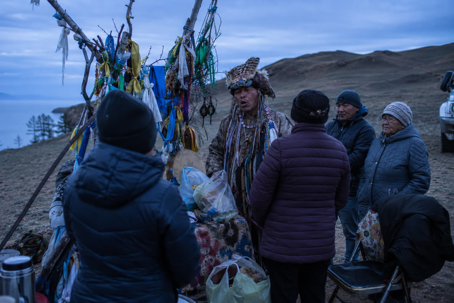 A shaman singing before a serge post hung with ribbons on a hillside above Lake Baikal, a small group standing with him at dusk